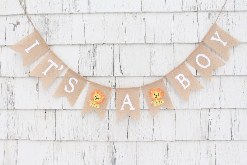 May include: A burlap banner with the words "It's a Boy" in white letters. The banner is decorated with small lion illustrations.