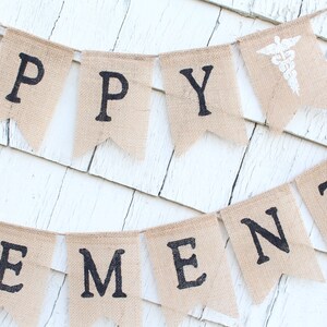 May include: A burlap banner with the words "Happy Retirement" in black letters. The banner is strung on a white wall and has a small white medical symbol on one of the flags.
