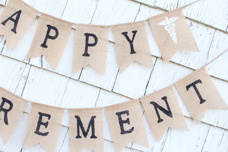 May include: A burlap banner with the words "Happy Retirement" in black letters. The banner is strung on a white wall and has a small white medical symbol on one of the flags.