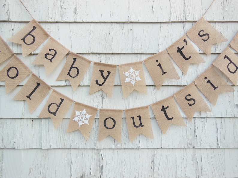 May include: A burlap banner with the text "Baby it's cold outside" in black lettering. The banner is decorated with white snowflakes.