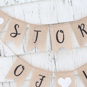 May include: A burlap banner with the words "Our Story" in black script. The banner is strung with white twine and has white felt hearts on either end.