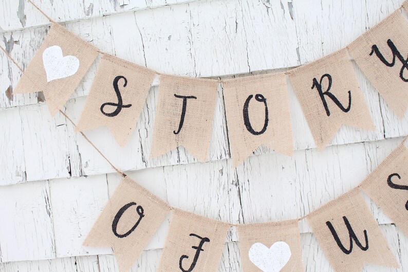May include: A burlap banner with the words "Our Story" in black script. The banner is strung with white twine and has white felt hearts on either end.