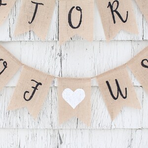 May include: A burlap banner with the words "Our Story" and a white heart in the centre. The banner is strung on a white wooden wall.