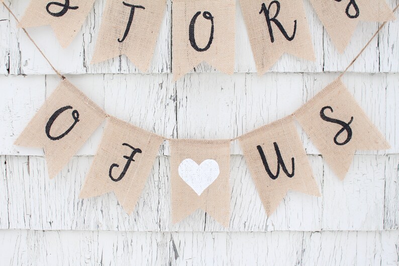 May include: A burlap banner with the words "Our Story" and a white heart in the centre. The banner is strung on a white wooden wall.