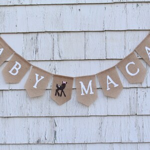 May include: A burlap banner with the words "BABY MACAY" in white letters. A small brown deer with a pink bow is between the "Y" and the "M".