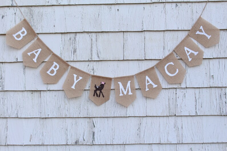 May include: A burlap banner with the words "BABY MACAY" in white letters. A small brown deer with a pink bow is between the "Y" and the "M".