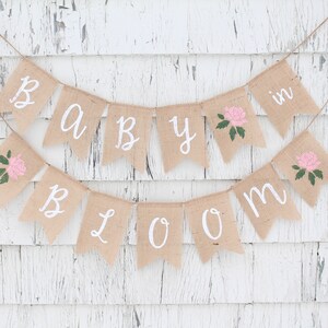 May include: A burlap banner with the words "Baby in Bloom" in white paint. The banner has pink flowers with green leaves on each flag.