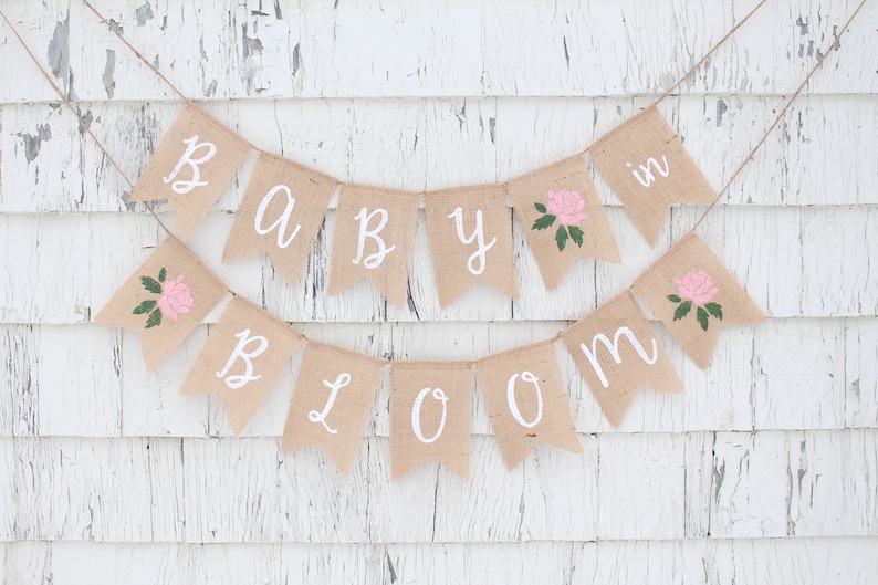 May include: A burlap banner with the words "Baby in Bloom" in white paint. The banner has pink flowers with green leaves on each flag.