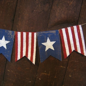 May include: A string of red, white, and blue bunting flags with white stars on a blue background. The flags are made of burlap and are strung together with twine. The flags are hanging against a dark wood background.