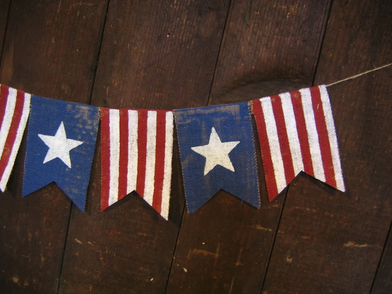 May include: A string of red, white, and blue bunting flags with white stars on a blue background. The flags are made of burlap and are strung together with twine. The flags are hanging against a dark wood background.