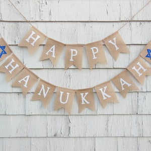 May include: A burlap banner with the words "HAPPY HANUKKAH" in white letters. The banner includes blue Star of David symbols. The banner is hanging against a white, weathered wood background.