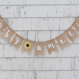 May include: A burlap banner with the words "baby Amelia" in white cursive lettering. A small yellow sunflower is on the "y" flag.