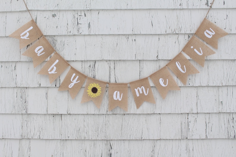 May include: A burlap banner with the words "baby Amelia" in white cursive lettering. A small yellow sunflower is on the "y" flag.