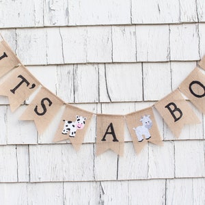 Puede incluir: Una pancarta de arpillera con las palabras "It's a Boy" en letras negras. La pancarta est&aacute; decorada con una vaca y una cabra.