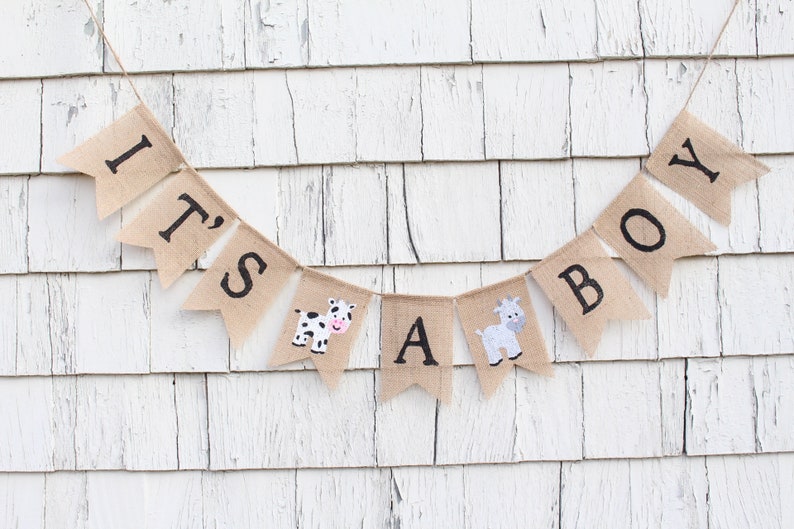Puede incluir: Una pancarta de arpillera con las palabras "It's a Boy" en letras negras. La pancarta est&aacute; decorada con una vaca y una cabra.