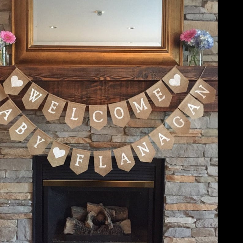 Puede incluir: Una pancarta de arpillera con letras blancas y corazones que dice "Bienvenido beb&eacute; Flanagan". La pancarta est&aacute; colgada sobre una chimenea.