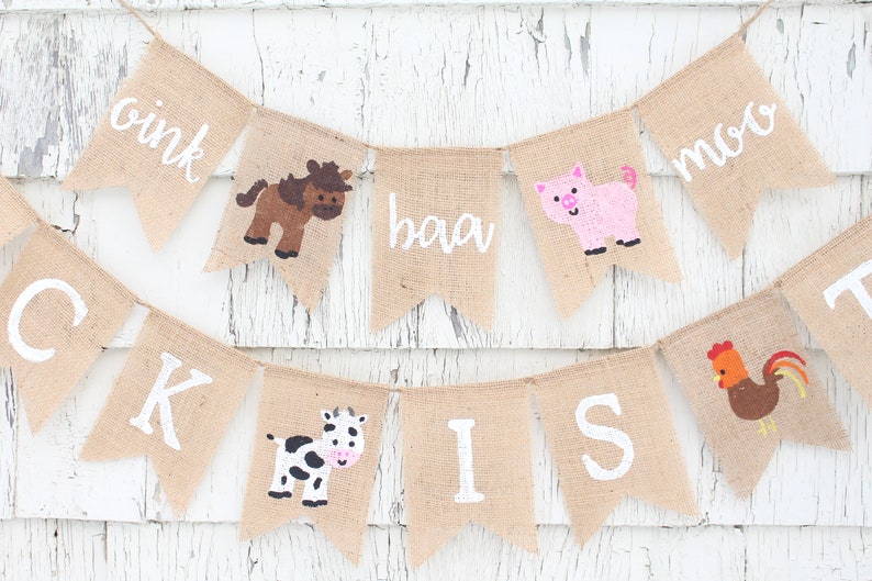 Pu&ograve; includere: Un banner di iuta con testo bianco e illustrazioni di animali della fattoria. Il banner include le parole "oink", "baa" e "moo", insieme a immagini di un cavallo, un maiale, una mucca e un gallo. Il banner &egrave; infilato su uno spago.