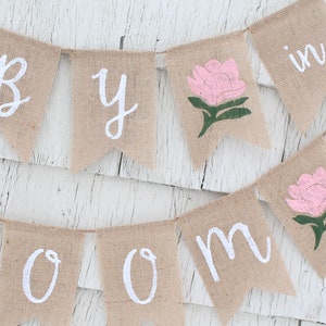 K&ouml;nnte beinhalten: Ein Banner aus Jute mit den Worten "Baby in Bloom" in wei&szlig;er Farbe. Das Banner ist mit rosa und gr&uuml;nen Blumen bemalt.