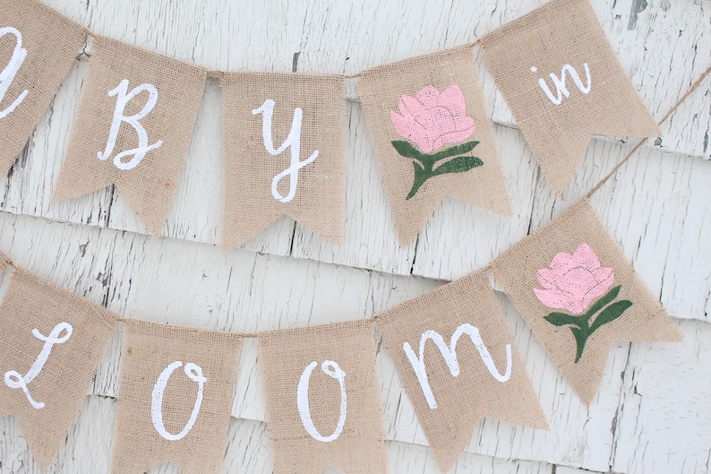 K&ouml;nnte beinhalten: Ein Banner aus Jute mit den Worten "Baby in Bloom" in wei&szlig;er Farbe. Das Banner ist mit rosa und gr&uuml;nen Blumen bemalt.