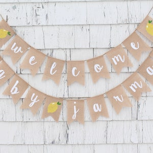 May include: A burlap banner with the words "Welcome Baby Jaman" in white lettering. The banner is decorated with small yellow lemons with green leaves.