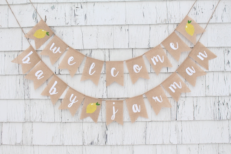 May include: A burlap banner with the words "Welcome Baby Jaman" in white lettering. The banner is decorated with small yellow lemons with green leaves.