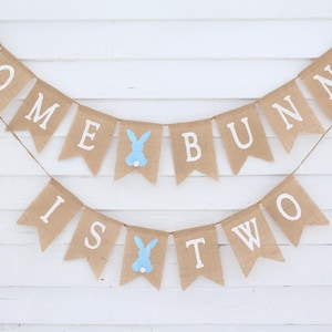 May include: A burlap banner with the text "Some Bunny Is Two" in white letters. The banner is decorated with blue bunny cutouts.