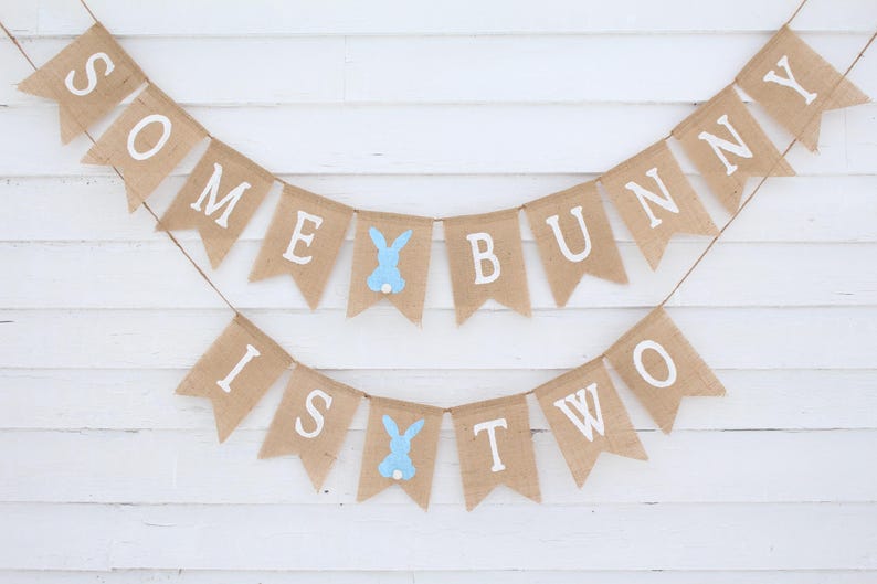 May include: A burlap banner with the text "Some Bunny Is Two" in white letters. The banner is decorated with blue bunny cutouts.