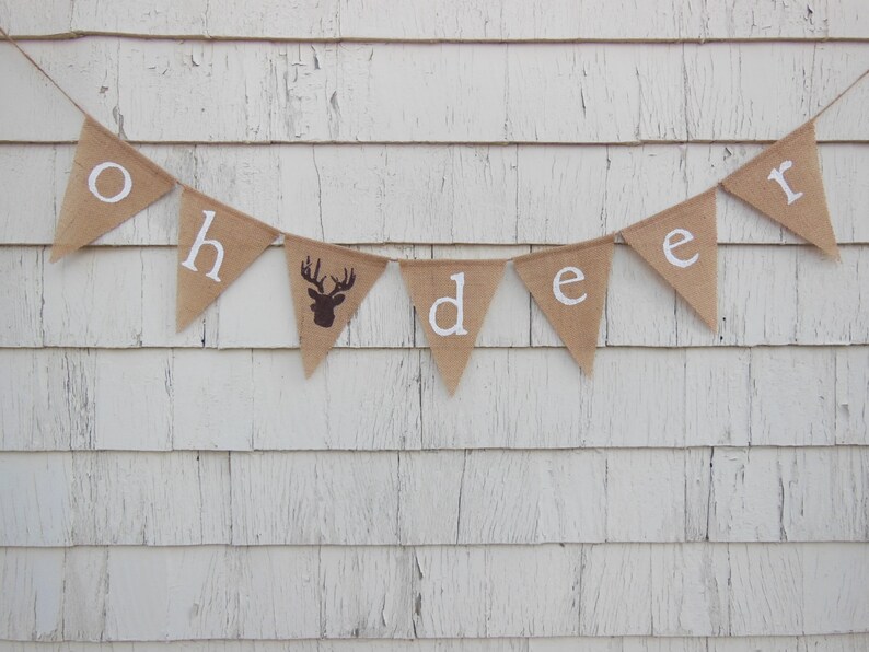 Puede incluir: Una pancarta de arpillera con las palabras "Oh Deer" en letras blancas. La pancarta est&aacute; colgada en una pared de madera blanca. Una silueta de ciervo marr&oacute;n se encuentra en el centro de la pancarta.
