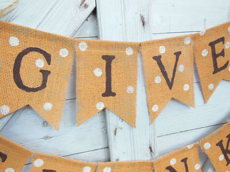 May include: A burlap banner with the word "GIVE" in dark brown letters. The orange pennants have white polka dots. The banner is strung on a natural twine and is against a white wooden background.