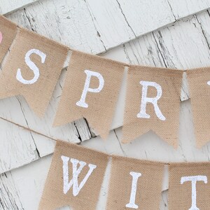 May include: A burlap banner with the words "Spring With" in white paint. A pink heart is on the left side of the banner.