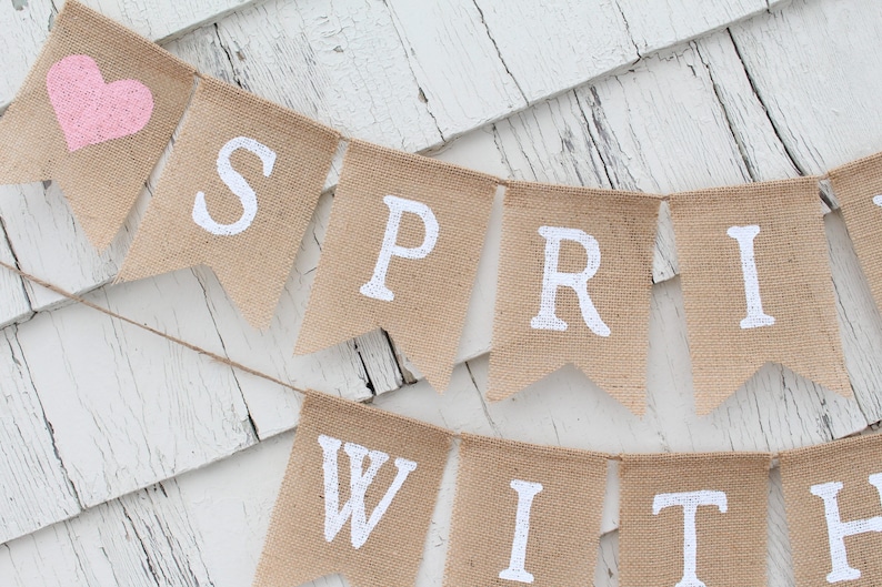 May include: A burlap banner with the words "Spring With" in white paint. A pink heart is on the left side of the banner.