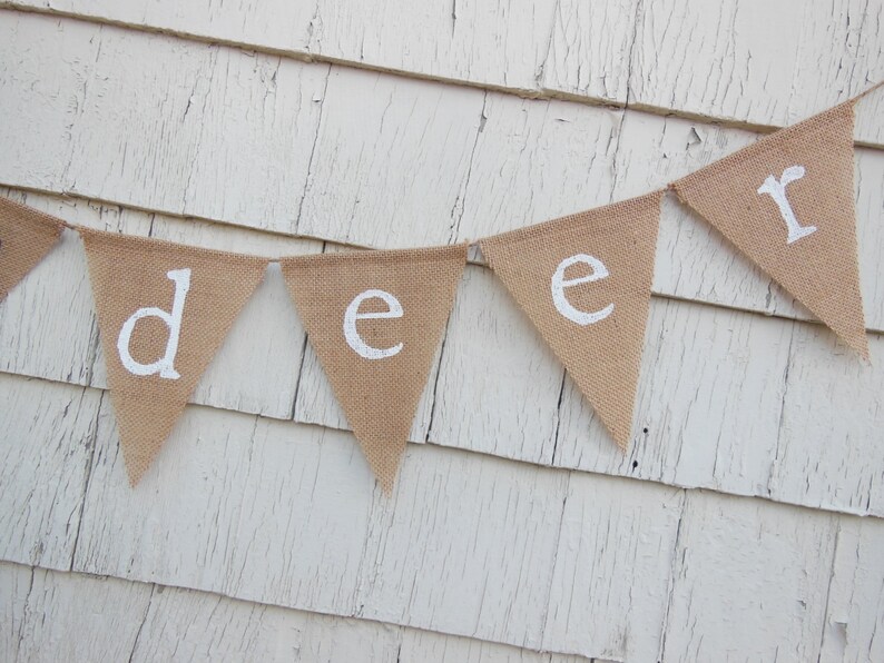 Puede incluir: Una pancarta de arpillera con la palabra "deer" escrita en pintura blanca. La pancarta est&aacute; colgada en una pared de madera blanca.