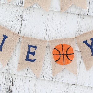 Pu&ograve; includere: Uno striscione di juta con la parola "BOY" scritta in lettere blu. Una palla da basket si trova tra le lettere "O" e "Y".