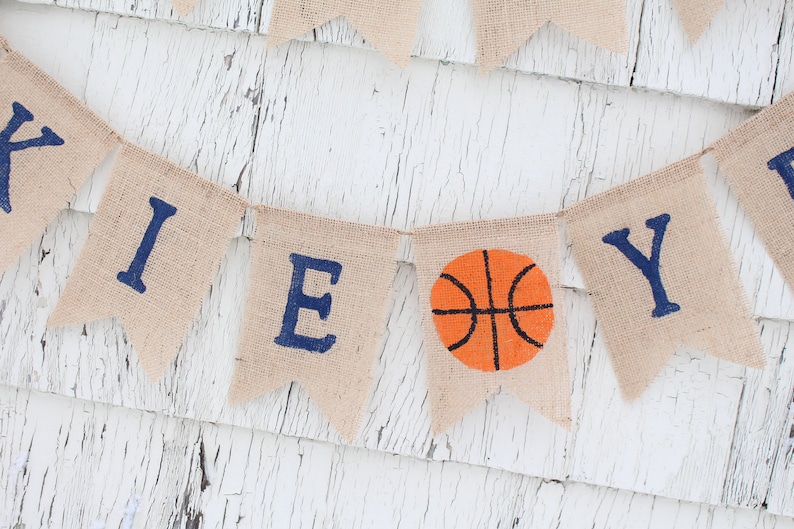 Pu&ograve; includere: Uno striscione di juta con la parola "BOY" scritta in lettere blu. Una palla da basket si trova tra le lettere "O" e "Y".