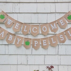 K&ouml;nnte beinhalten: Ein Wimpelbanner aus Jute mit den Worten "WELCOME BABY PELSIS" in wei&szlig;en Buchstaben. Das Banner hat einen rustikalen Look mit gr&uuml;nen Apfel-Illustrationen auf einigen Wimpeln. Es ist an einer Schnur befestigt und h&auml;ngt vor einem wei&szlig;en Holzgrund.