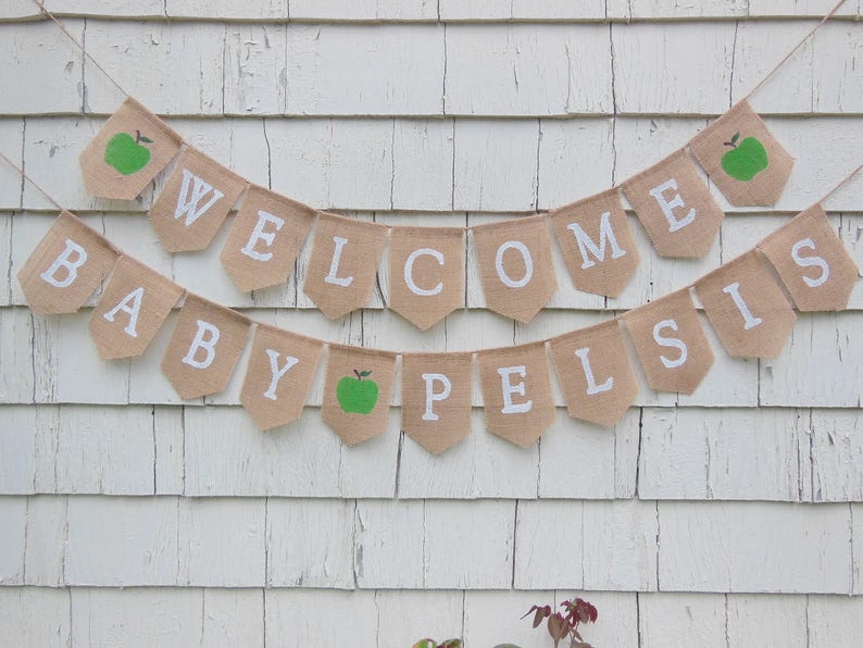 K&ouml;nnte beinhalten: Ein Wimpelbanner aus Jute mit den Worten "WELCOME BABY PELSIS" in wei&szlig;en Buchstaben. Das Banner hat einen rustikalen Look mit gr&uuml;nen Apfel-Illustrationen auf einigen Wimpeln. Es ist an einer Schnur befestigt und h&auml;ngt vor einem wei&szlig;en Holzgrund.