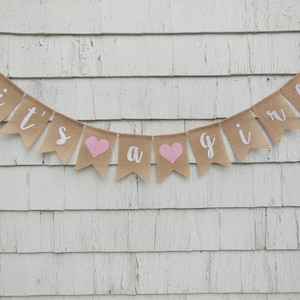 May include: A burlap banner with the words "it's a girl" in white lettering and pink hearts. The banner is hanging on a white wooden wall.