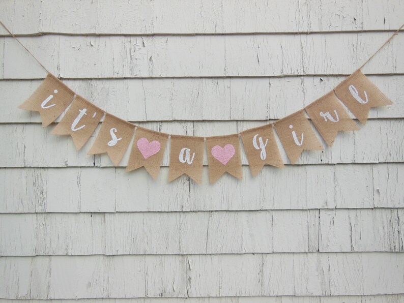 May include: A burlap banner with the words "it's a girl" in white lettering and pink hearts. The banner is hanging on a white wooden wall.