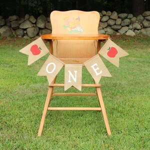 May include: A burlap banner with red apples and the word "ONE" in white letters hangs on a wooden high chair. The chair is in front of a stone wall and green grass.