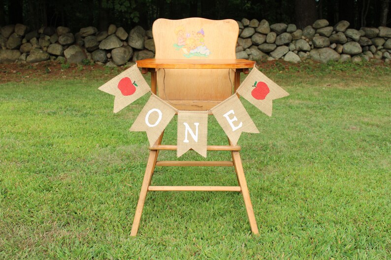 May include: A burlap banner with red apples and the word "ONE" in white letters hangs on a wooden high chair. The chair is in front of a stone wall and green grass.