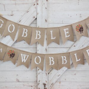 May include: Thanksgiving banner made of burlap with the words "GOBBLE TILL YA WOBBLE" in white stencil letters. Each pennant is tan with a turkey graphic.