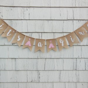 May include: A burlap banner with the text "it's a girl" in white lettering. The banner is decorated with pink and white teepees.