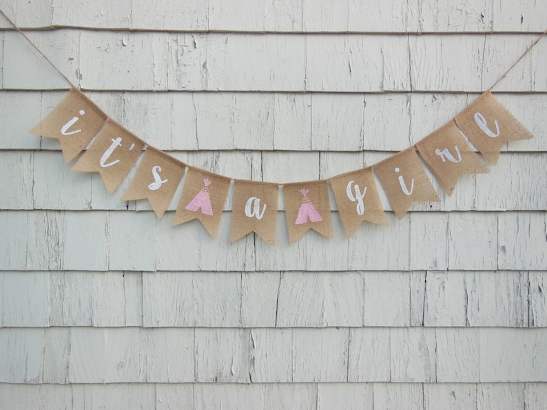 May include: A burlap banner with the text "it's a girl" in white lettering. The banner is decorated with pink and white teepees.