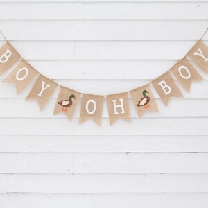 May include: A burlap banner with the words "Oh Boy" and two ducks, hanging on a white wood slat wall. The banner is strung with twine.