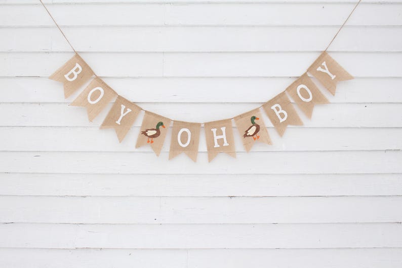 May include: A burlap banner with the words "Oh Boy" and two ducks, hanging on a white wood slat wall. The banner is strung with twine.