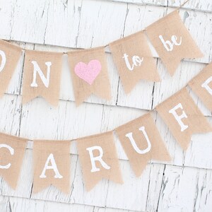 May include: A burlap banner with the words "Soon to be" and a pink heart. The banner is strung with white twine and has white letters.