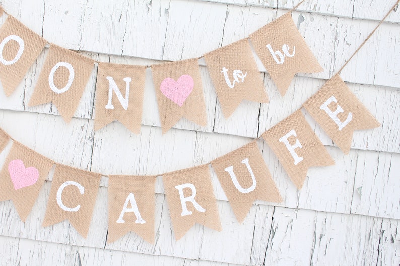 May include: A burlap banner with the words "Soon to be" and a pink heart. The banner is strung with white twine and has white letters.