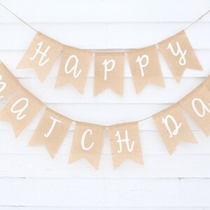 May include: A banner with the words "Happy Match Day" written in white script on individual tan pennants. The pennants are strung on a thin twine against a white wooden background.