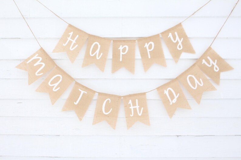 May include: A banner with the words "Happy Match Day" written in white script on individual tan pennants. The pennants are strung on a thin twine against a white wooden background.
