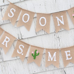 May include: A burlap banner with the words "Soon to be Mrs." and a cactus design. The banner is strung on a white wooden wall.